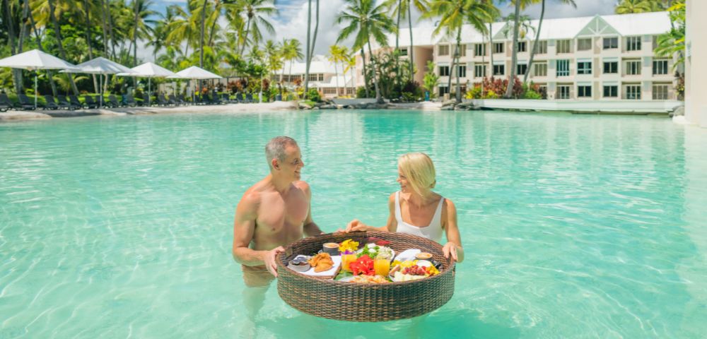 A couple stands in a shallow, clear turquoise pool, smiling at each other and holding a large, round wicker tray laden with a colorful breakfast, including fruit, pastries, and drinks. In the background, palm trees line a sandy beach with white umbrellas and a resort building.