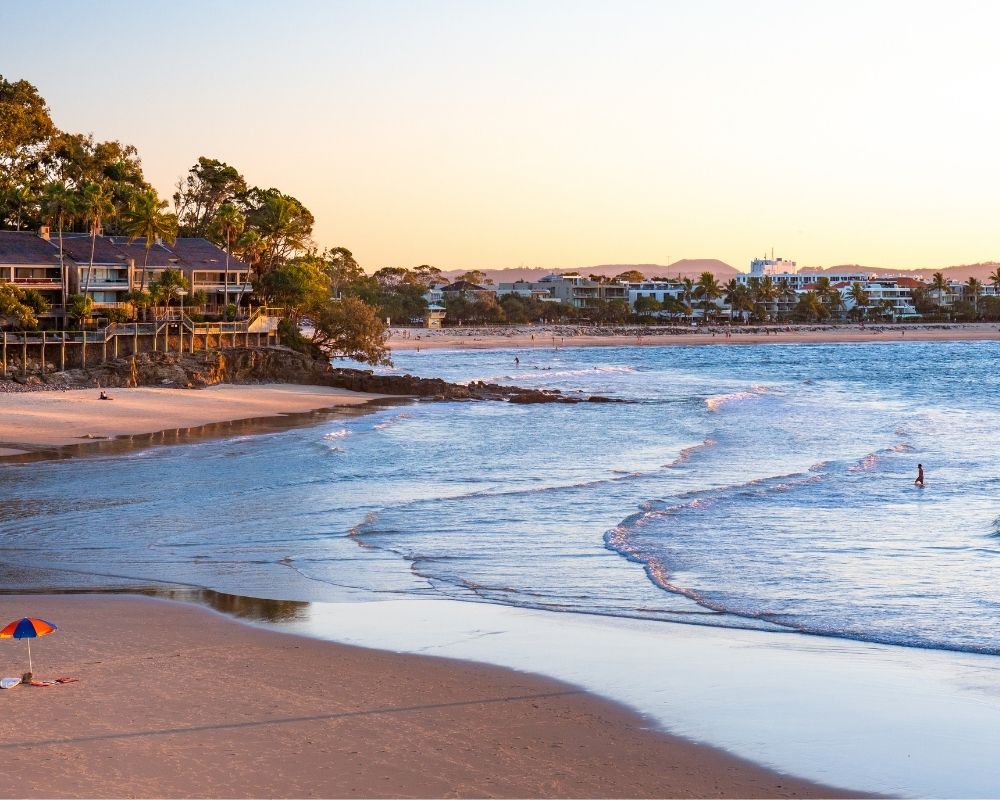 A wide sandy beach with gentle waves under a warm, hazy sky. A cluster of modern buildings with wooden balconies is nestled into a tree-covered bluff on the left. In the distance, a town stretches along the coast. Two individuals are in the shallow water, and a colorful umbrella stands on the sand.