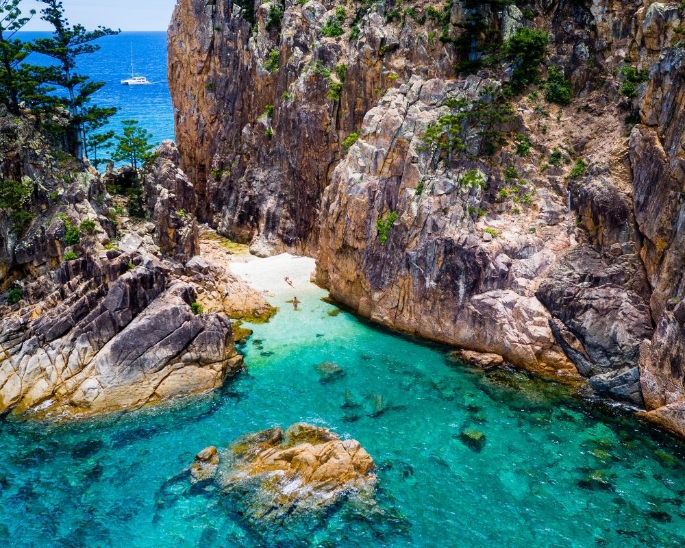 Aerial view of a secluded cove with crystal clear turquoise water, rocky cliffs, and a small sandy beach where two people are swimming.