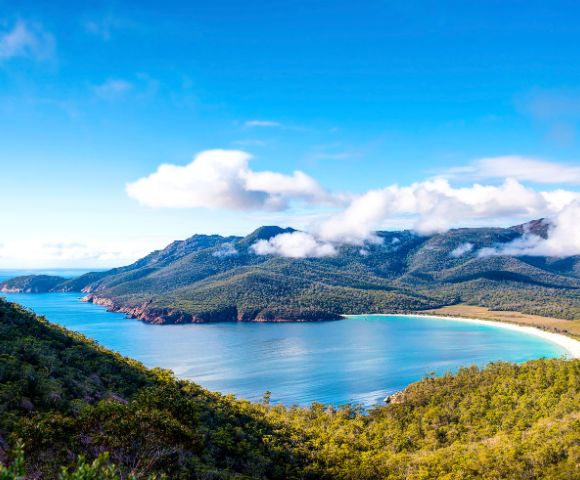 Panoramic view of a horseshoe-shaped bay with turquoise water, white sandy beach, and tree-covered mountains under a bright blue sky with scattered white clouds.