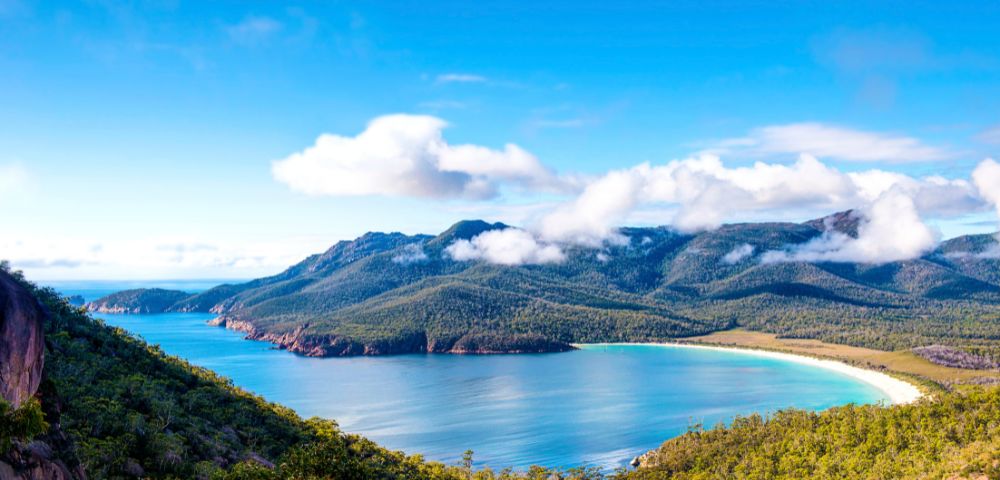 Panoramic view of a horseshoe-shaped bay with turquoise water, white sandy beach, and tree-covered mountains under a bright blue sky with scattered white clouds.
