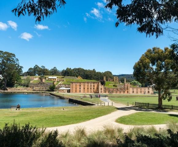 A historic convict settlement with sandstone buildings is situated beside a calm body of water under a clear blue sky, with grassy fields and trees in the foreground.