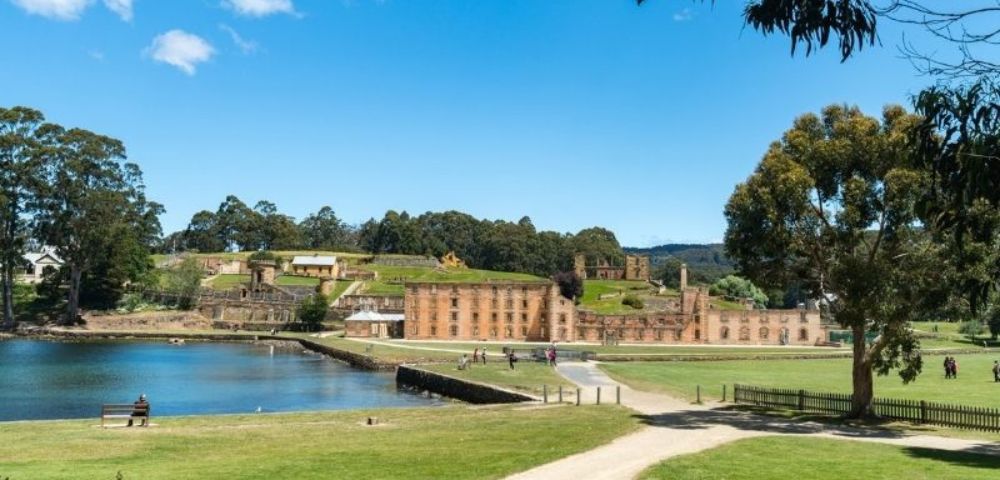 A historic convict settlement with sandstone buildings is situated beside a calm body of water under a clear blue sky, with grassy fields and trees in the foreground.