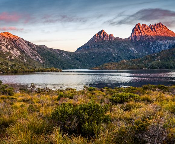 Sunrise illuminates jagged mountain peaks reflecting in a calm lake surrounded by dense forest and a foreground of golden marsh grass and low-lying shrubs.
