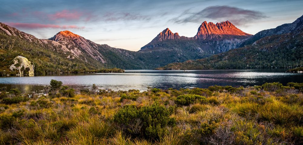 Sunrise illuminates jagged mountain peaks reflecting in a calm lake surrounded by dense forest and a foreground of golden marsh grass and low-lying shrubs.