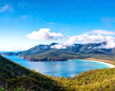 Panoramic view of a horseshoe-shaped bay with turquoise water, white sandy beach, and tree-covered mountains under a bright blue sky with scattered white clouds.