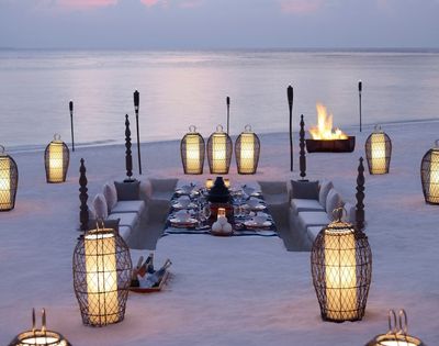 An elegant outdoor dining setup on a sandy beach at dusk, featuring a sunken seating area with plush cushions surrounding a beautifully set table with white tableware. Illuminated by ornate lanterns and torches, with a crackling bonfire in the background.