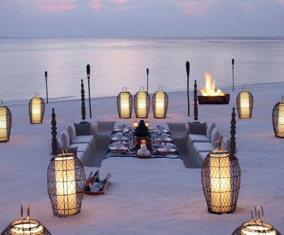 An elegant outdoor dining setup on a sandy beach at dusk, featuring a sunken seating area with plush cushions surrounding a beautifully set table with white tableware. Illuminated by ornate lanterns and torches, with a crackling bonfire in the background.