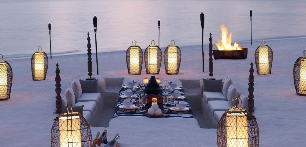 An elegant outdoor dining setup on a sandy beach at dusk, featuring a sunken seating area with plush cushions surrounding a beautifully set table with white tableware. Illuminated by ornate lanterns and torches, with a crackling bonfire in the background.