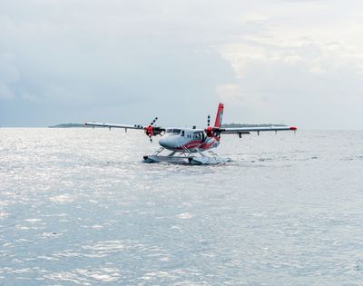 A twin-engine seaplane with red and white markings rests on calm water, with a distant island visible under an overcast sky.