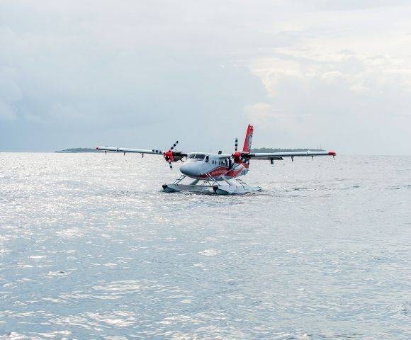 A twin-engine seaplane with red and white markings rests on calm water, with a distant island visible under an overcast sky.