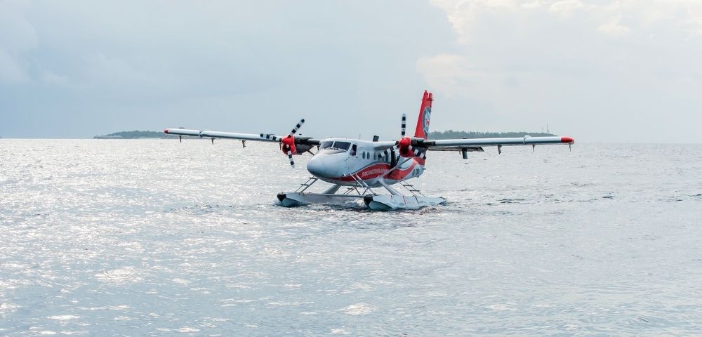 A twin-engine seaplane with red and white markings rests on calm water, with a distant island visible under an overcast sky.