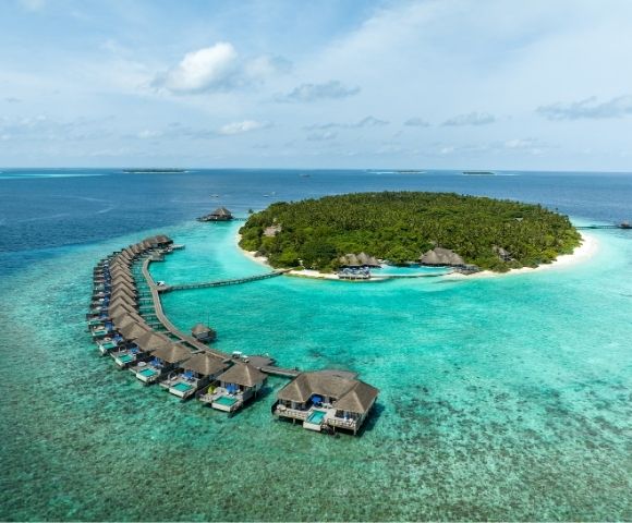 Aerial view of a tropical island resort with overwater bungalows curving into a turquoise lagoon and a lush green island in the background.