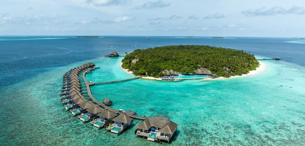 Aerial view of a tropical island resort with overwater bungalows curving into a turquoise lagoon and a lush green island in the background.