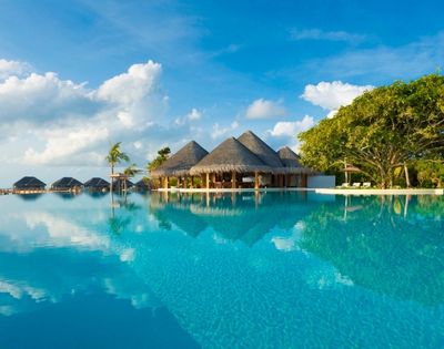 A tranquil infinity pool with a thatched-roof resort building and several overwater bungalows in the background, set against a backdrop of blue sky with fluffy clouds and lush green trees.