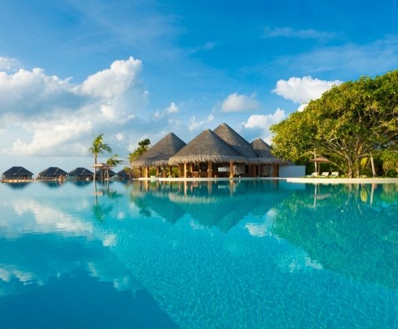A tranquil infinity pool with a thatched-roof resort building and several overwater bungalows in the background, set against a backdrop of blue sky with fluffy clouds and lush green trees.