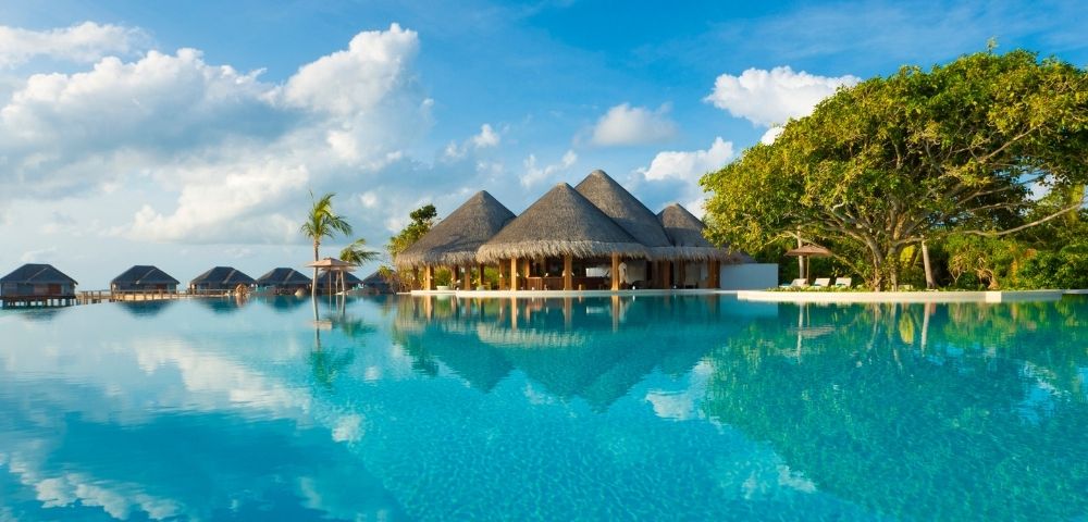 A tranquil infinity pool with a thatched-roof resort building and several overwater bungalows in the background, set against a backdrop of blue sky with fluffy clouds and lush green trees.