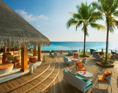 A tropical seaside lounge area with wooden decking, comfortable seating, palm trees, and a clear blue ocean in the background.