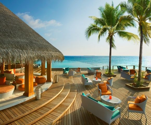 A tropical seaside lounge area with wooden decking, comfortable seating, palm trees, and a clear blue ocean in the background.