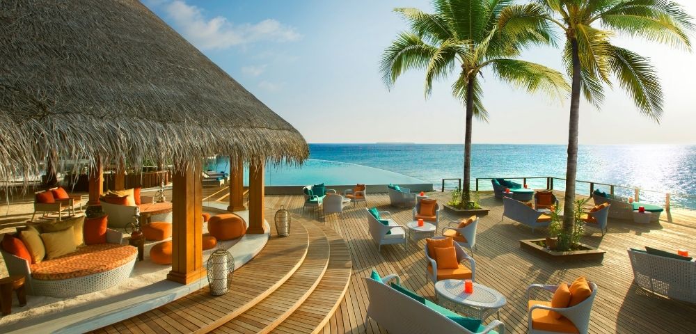A tropical seaside lounge area with wooden decking, comfortable seating, palm trees, and a clear blue ocean in the background.
