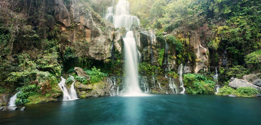 A serene multi-tiered waterfall cascades over mossy cliffs into a deep turquoise pool, surrounded by lush green forest and misty light.