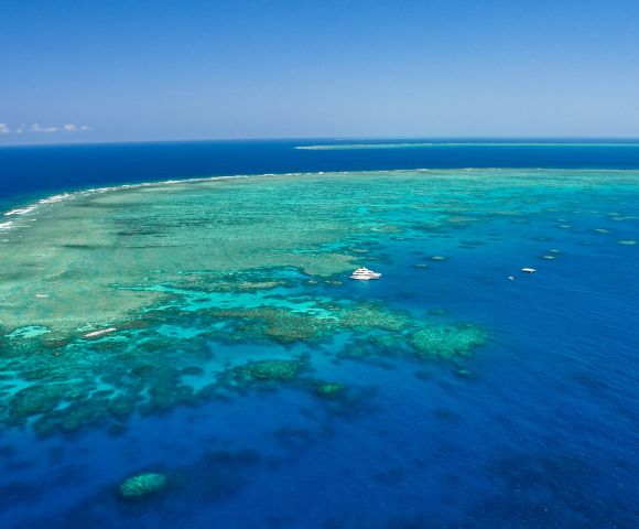 Aerial view of a vibrant coral reef system in clear turquoise and deep blue ocean waters, with a large white yacht anchored nearby.