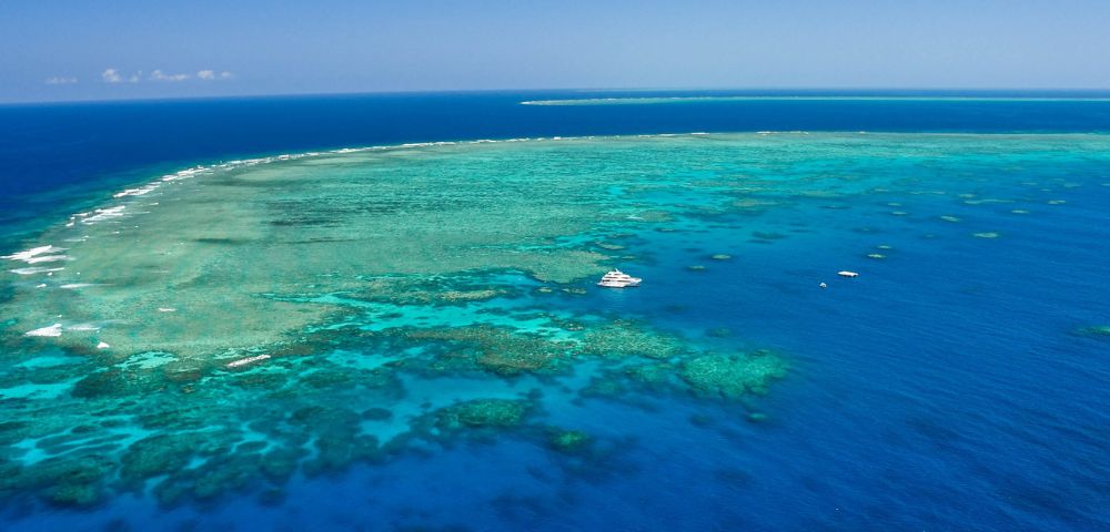 Aerial view of a vibrant coral reef system in clear turquoise and deep blue ocean waters, with a large white yacht anchored nearby.