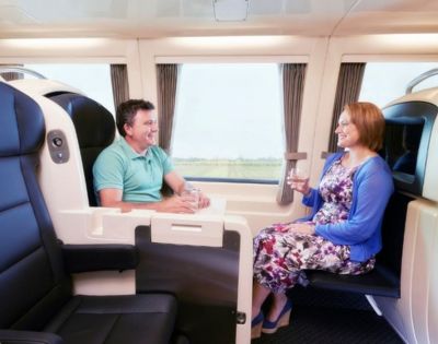 Two passengers enjoy drinks in first-class train seating with large windows and comfortable seats.