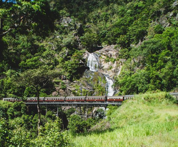A vintage train with red and white carriages crosses a viaduct in front of a cascading waterfall on a lush green mountainside.