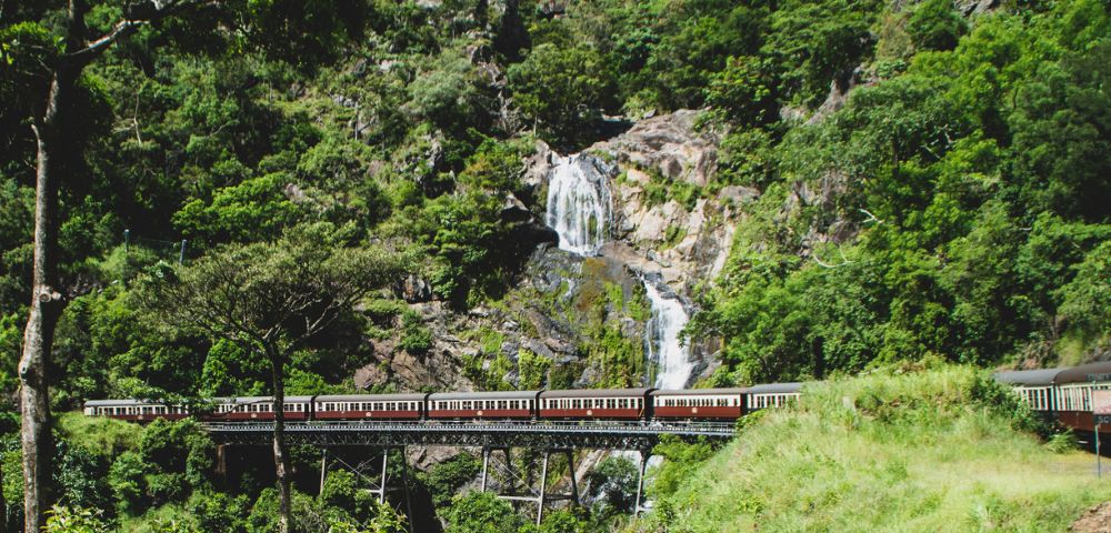A vintage train with red and white carriages crosses a viaduct in front of a cascading waterfall on a lush green mountainside.