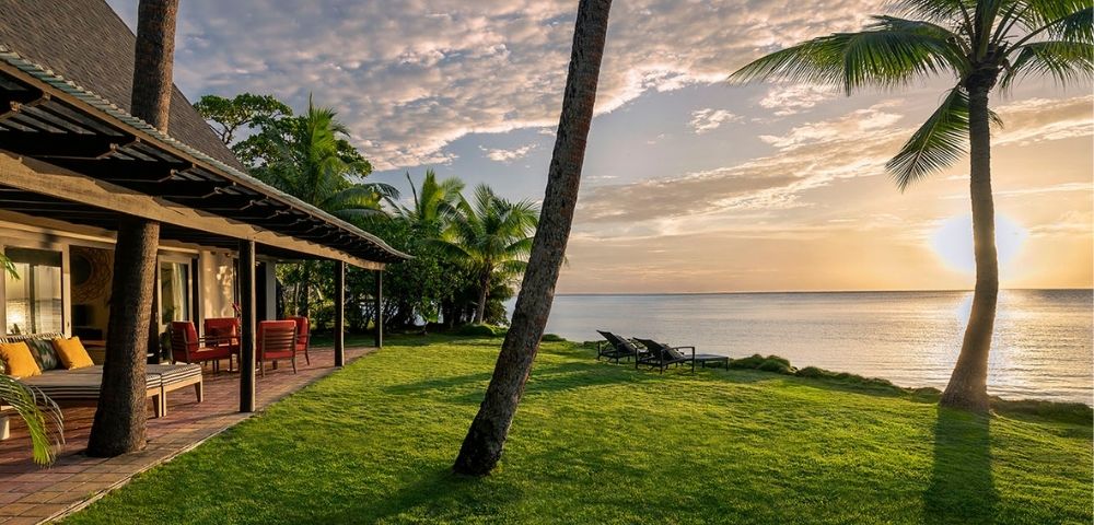 Tropical bungalow with ocean view at sunset, featuring a covered porch with seating and loungers on a grassy lawn.