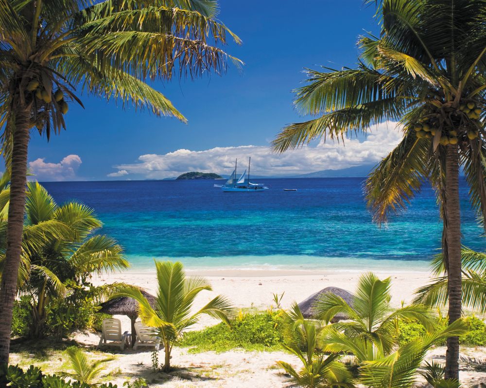 A bright blue sky with fluffy white clouds sits above a calm, turquoise ocean, framed by lush green palm trees. In the distance, a sailboat with white sails glides across the water near a small, tropical island. White sand lines the shore, and thatched-roof umbrellas dot the beach.