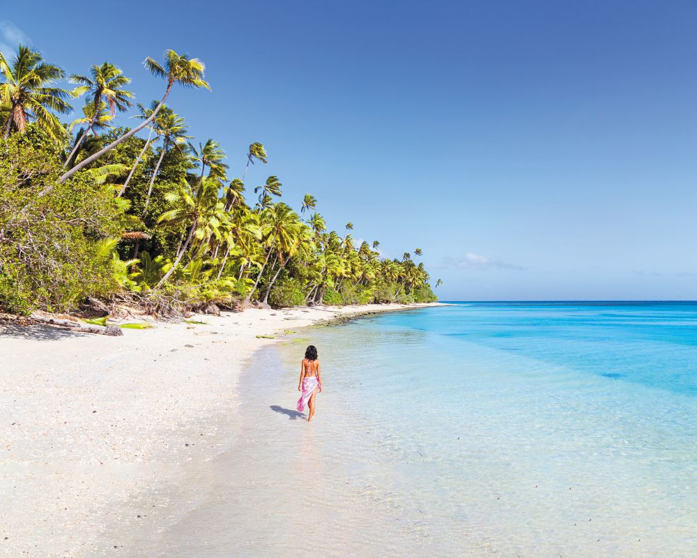 A person wearing a pink sarong walks on a pristine white sand beach lined with lush green palm trees and turquoise waters under a clear blue sky.