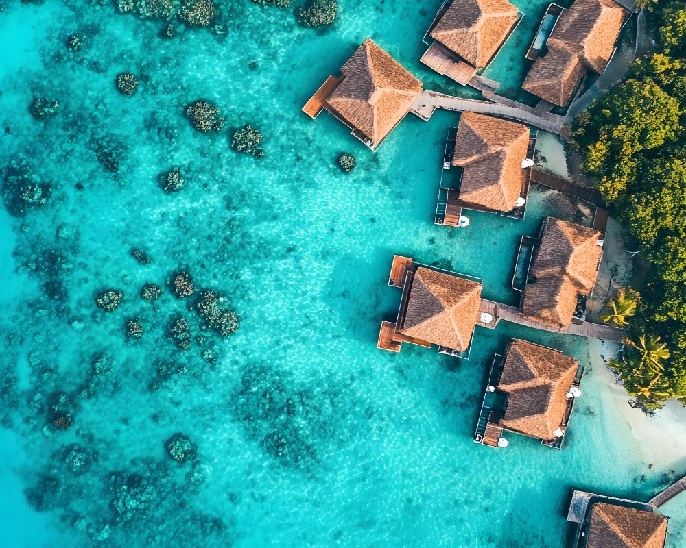 Aerial view of overwater bungalows with thatched roofs nestled in a crystal-clear turquoise lagoon dotted with coral reefs and surrounded by lush green foliage.