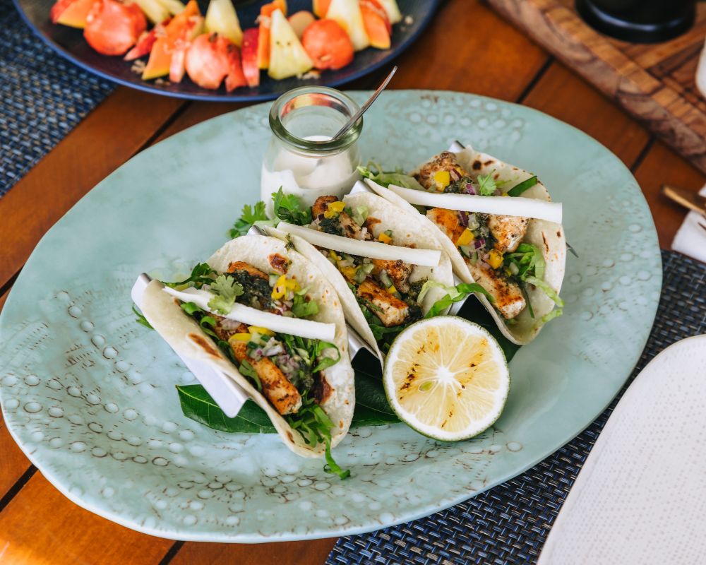 Three grilled chicken tacos filled with lettuce, corn, and a creamy sauce, served on a light blue patterned plate with a halved lime and a small dish of dipping sauce.