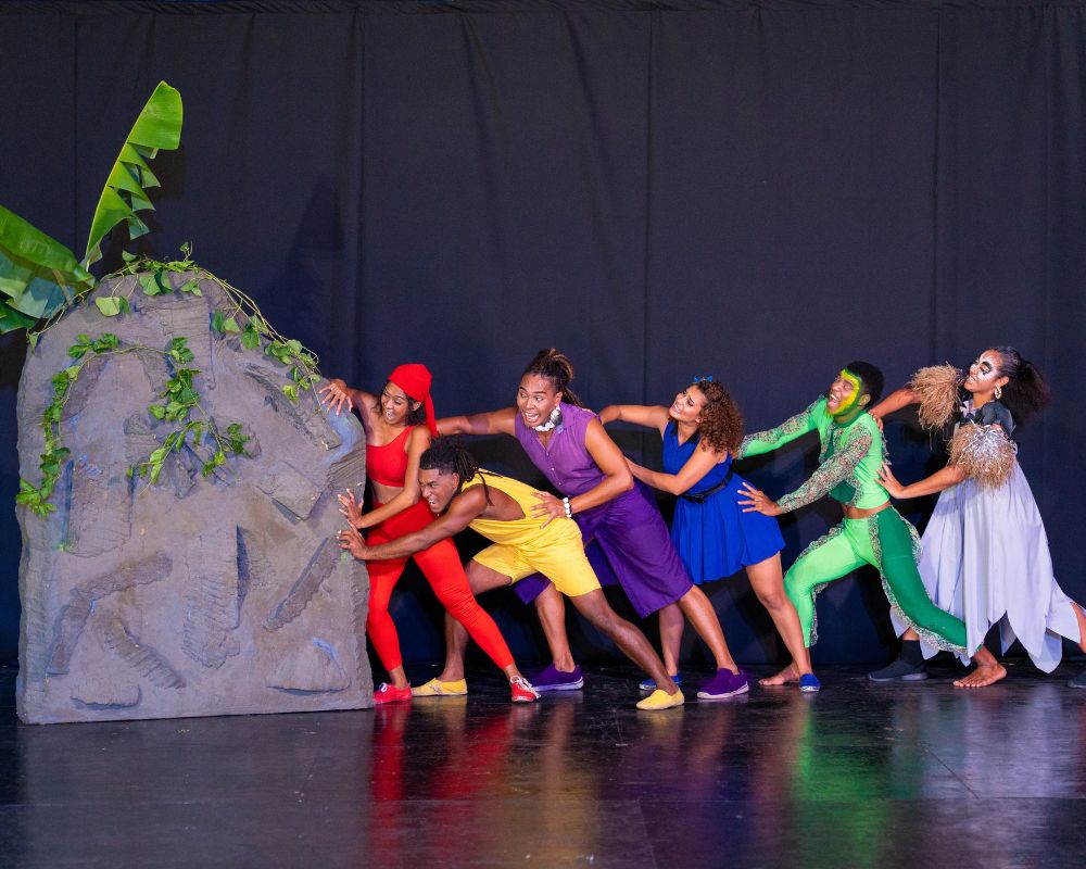 A diverse group of performers in brightly colored costumes push a large, gray, rock-like prop on a stage with a dark backdrop.
