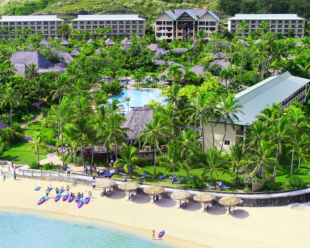 Aerial view of a tropical resort with multiple buildings, thatched-roof huts, lush green palm trees, a large swimming pool, and a sandy beach with kayaks lined up.