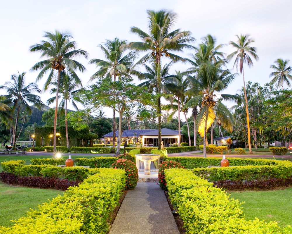 A pathway leads through manicured green hedges and flowerbeds towards a building with a dark roof, surrounded by numerous tall palm trees, with a fountain in the foreground and subtle lighting illuminating the scene at dusk.