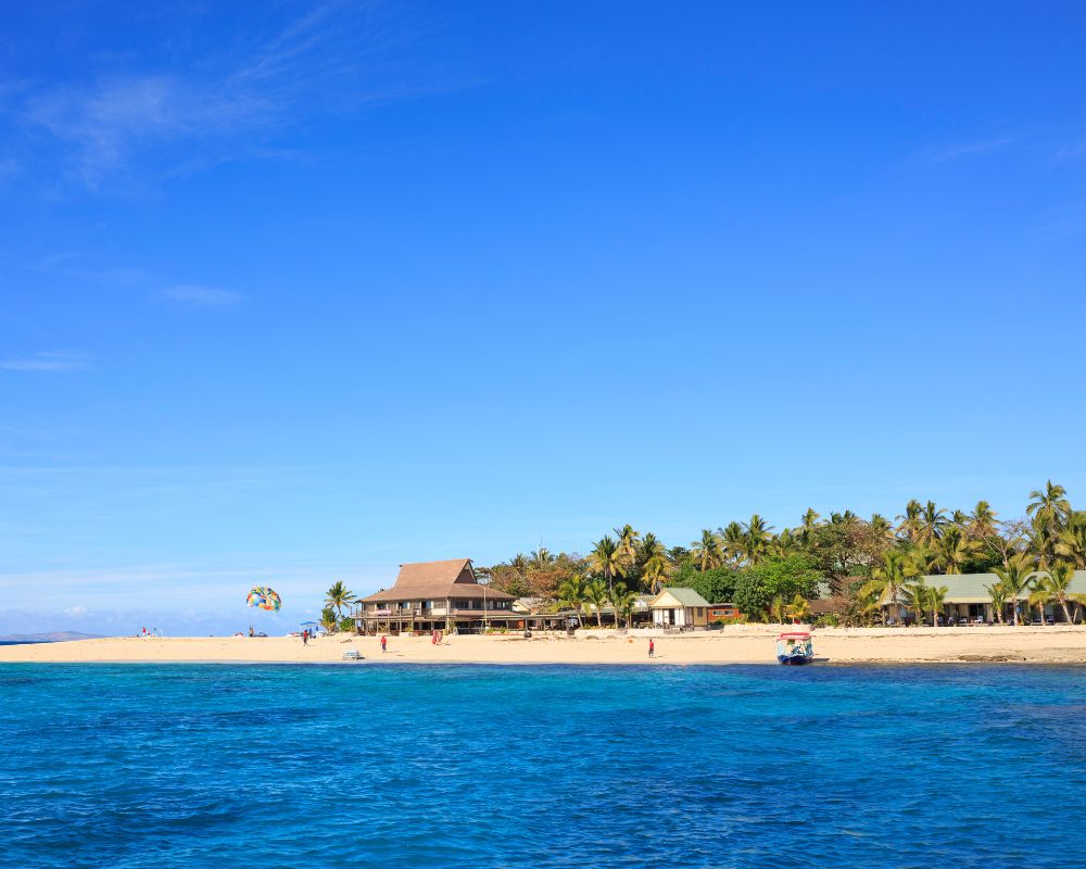 Bright blue ocean with clear sky over a tropical island beach featuring palm trees, resort buildings, and parasailing activity.