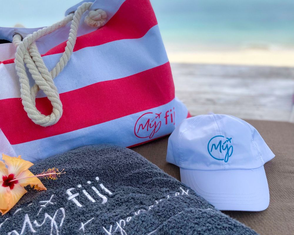 A beach bag with red and white stripes, a white baseball cap with a blue logo, and a dark gray towel with white embroidery and a yellow hibiscus flower, all placed on a lounge chair with the ocean in the background.
