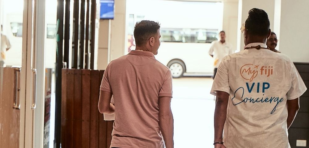 A man in a pink polo shirt and black shorts walks with a rolling suitcase, accompanied by a VIP Concierge employee wearing a white shirt with 
