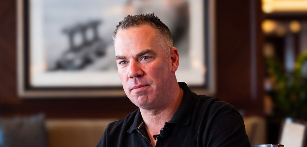 A man with short, graying hair and blue eyes, wearing a black polo shirt, sits in a chair in a softly lit room. Behind him, a large framed black and white photograph hangs on a dark wood paneled wall. The background is blurred, showing hints of seating and decorative lighting.
