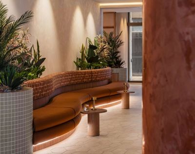 A curved, burnt orange upholstered sofa with a patterned backrest curves around a planter filled with lush green and pink foliage. Small, round wooden side tables with brass lamps and drinks are placed in front of the sofa, and soft LED lighting illuminates the base of the sofa, creating a warm and inviting atmosphere in a modern lounge or reception area.