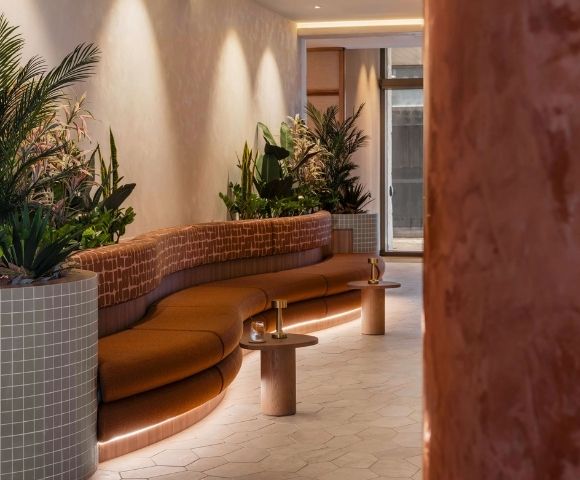 A curved, burnt orange upholstered sofa with a patterned backrest curves around a planter filled with lush green and pink foliage. Small, round wooden side tables with brass lamps and drinks are placed in front of the sofa, and soft LED lighting illuminates the base of the sofa, creating a warm and inviting atmosphere in a modern lounge or reception area.
