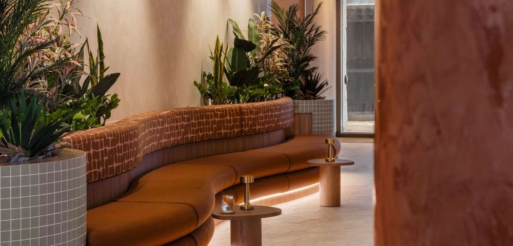 A curved, burnt orange upholstered sofa with a patterned backrest curves around a planter filled with lush green and pink foliage. Small, round wooden side tables with brass lamps and drinks are placed in front of the sofa, and soft LED lighting illuminates the base of the sofa, creating a warm and inviting atmosphere in a modern lounge or reception area.