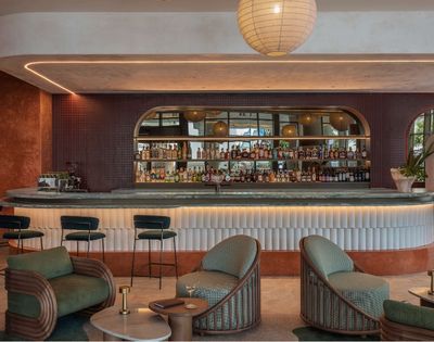 Modern bar with a curved counter featuring a stone top, shelves of liquor bottles, and bar stools, illuminated by soft accent lighting and a large pendant lamp overhead.