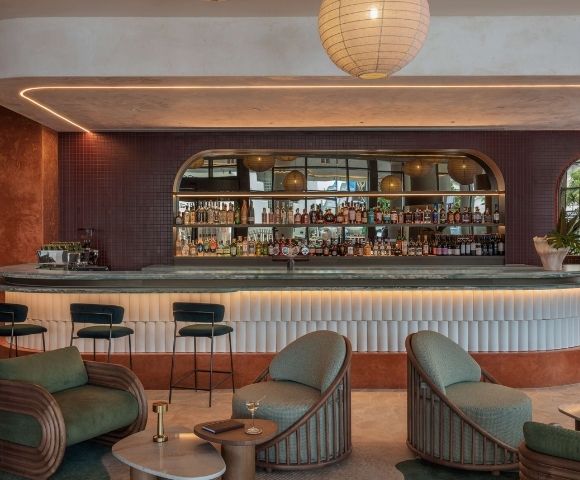 Modern bar with a curved counter featuring a stone top, shelves of liquor bottles, and bar stools, illuminated by soft accent lighting and a large pendant lamp overhead.