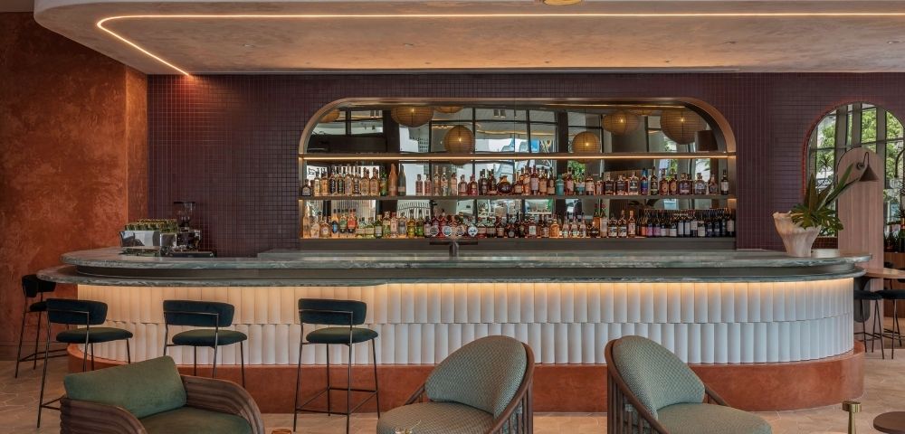 Modern bar with a curved counter featuring a stone top, shelves of liquor bottles, and bar stools, illuminated by soft accent lighting and a large pendant lamp overhead.