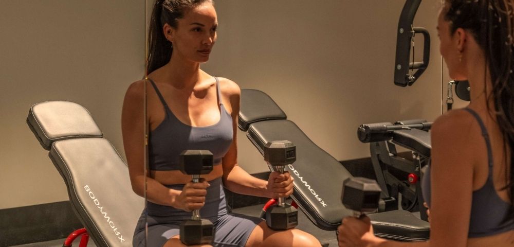 A woman in workout attire holds two dumbbells in a gym, reflected in a mirror.