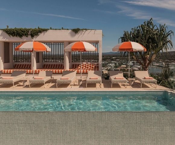 A person rests in a turquoise swimming pool with a tiled edge, overlooking a sunny rooftop lounge area with beige lounge chairs and orange and white striped umbrellas.